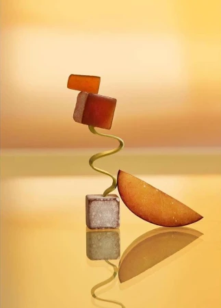 Still life photography featuring stacked cube-shaped fruit jellies and a citrus slice, balanced on a spiraling metal stand against a gradient orange background.