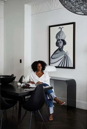 An adult female sits at a black dining table in a bright room, with artwork visible on the wall and natural light coming in.