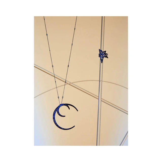 Elegant jewelry still life: A blue crescent moon necklace dangles next to a blue star-shaped ring, displayed on a clear stand against a peach background.