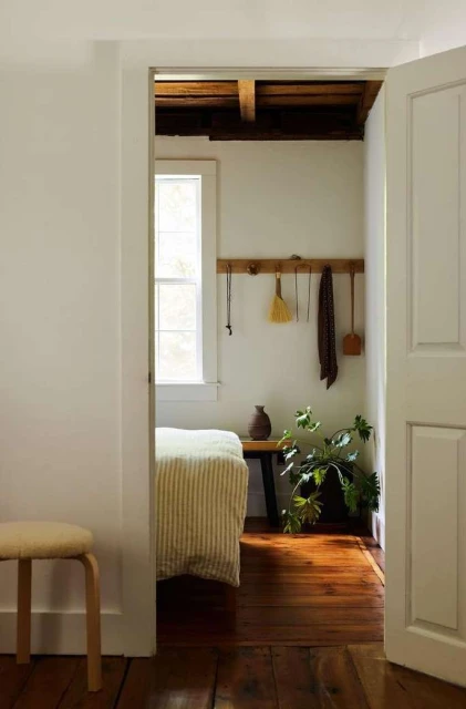 View of interior with white walls, wooden floors, and a peek into an adjacent room featuring a bed, plant, and various household items.