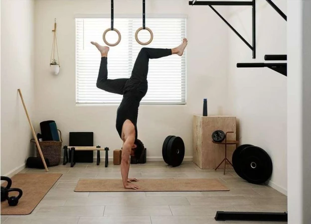 Adult male in black sportswear performs a handstand in a home gym with exercise equipment and a wood floor in a brightly lit room.