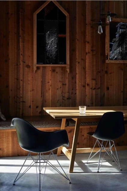 Interior view features a wooden-paneled wall, dining table with a glass, and black modern chairs under pendant lights. 