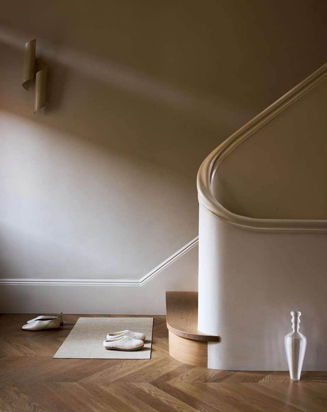 Minimalist interior design featuring a staircase with white banister, wooden floor, and light-colored slippers and vase, creating a serene ambiance.