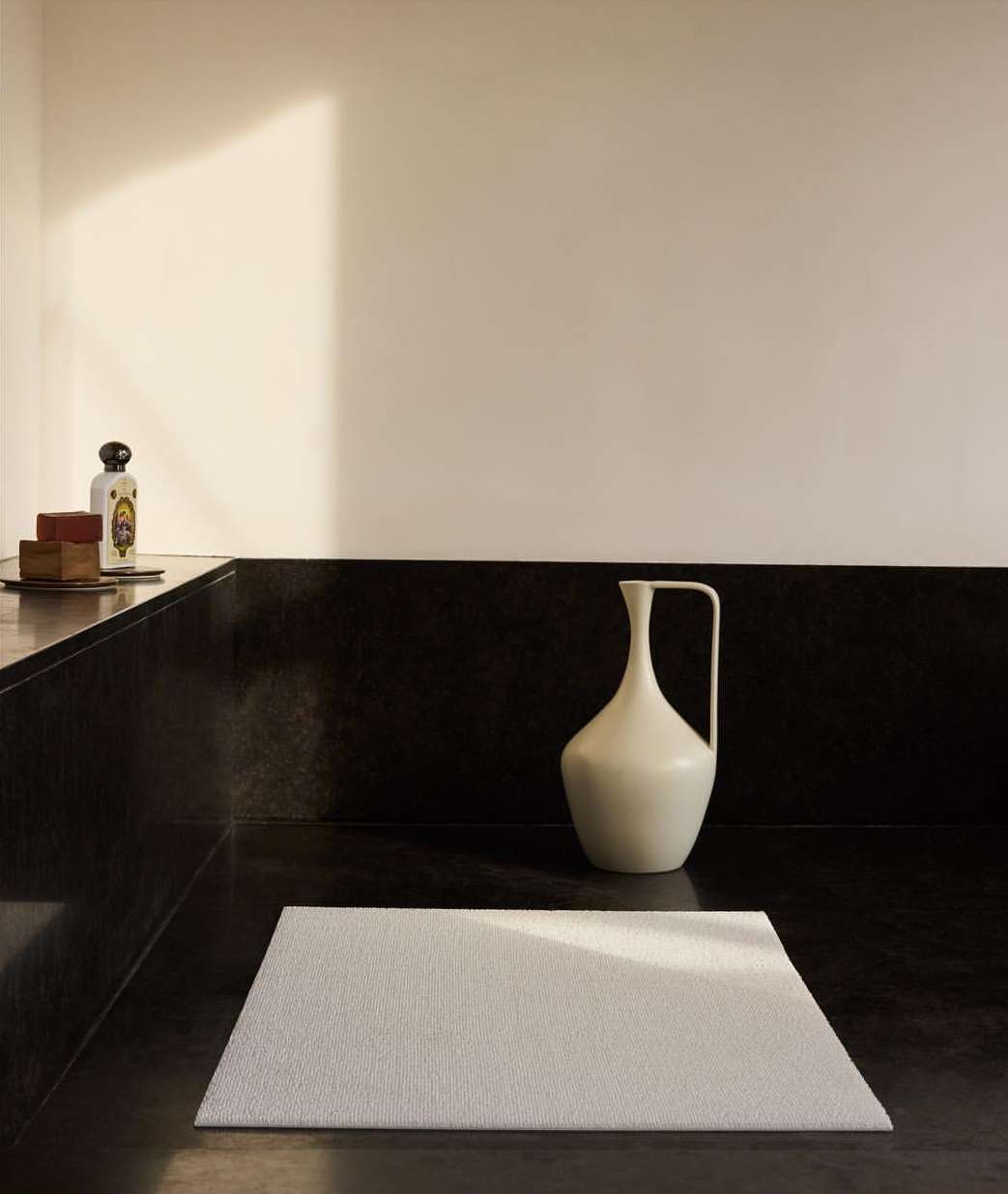 Minimalist interior shot of a white bath mat on a dark stone floor with a white vase and toiletry items in the background.