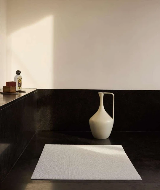 Minimalist interior shot of a white bath mat on a dark stone floor with a white vase and toiletry items in the background.