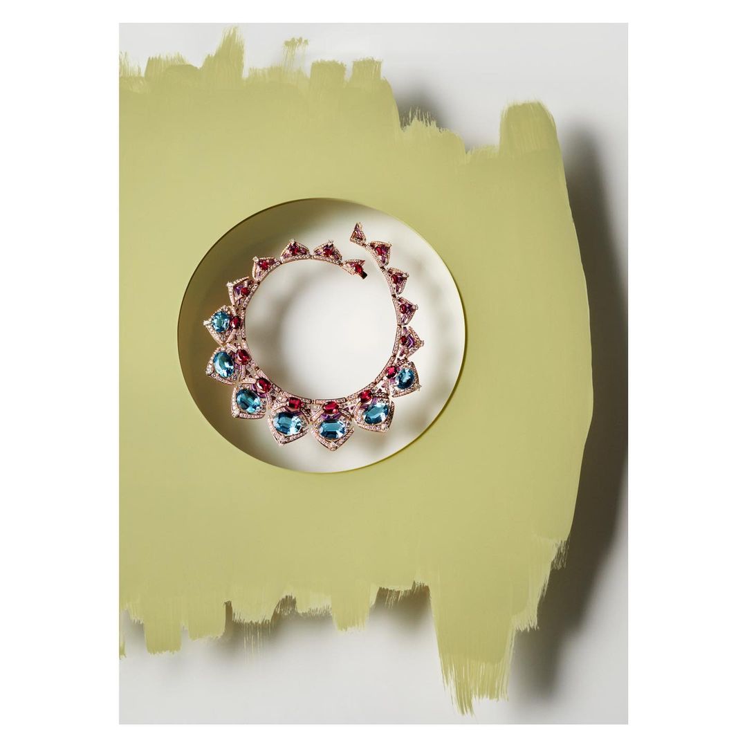 Rose gold necklace with blue and ruby gemstones on a painted surface, presented against a white backdrop for a modern, artistic jewelry display.