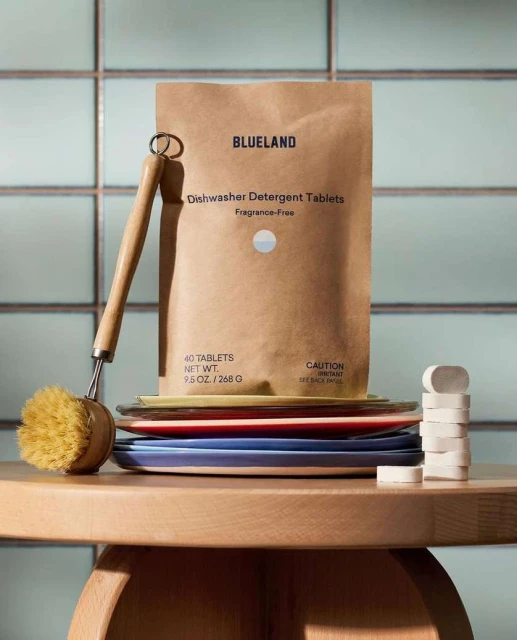 Blueland dishwasher detergent tablets in a brown paper bag next to a dish brush and stack of dishware on a light wood table.