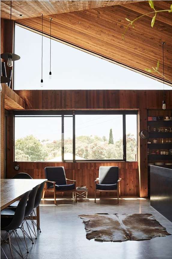 Living room interior with wood paneling, large windows overlooking trees, cowhide rug on the floor, two armchairs, and dining table.