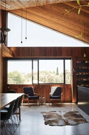 Living room interior with wood paneling, large windows overlooking trees, cowhide rug on the floor, two armchairs, and dining table.