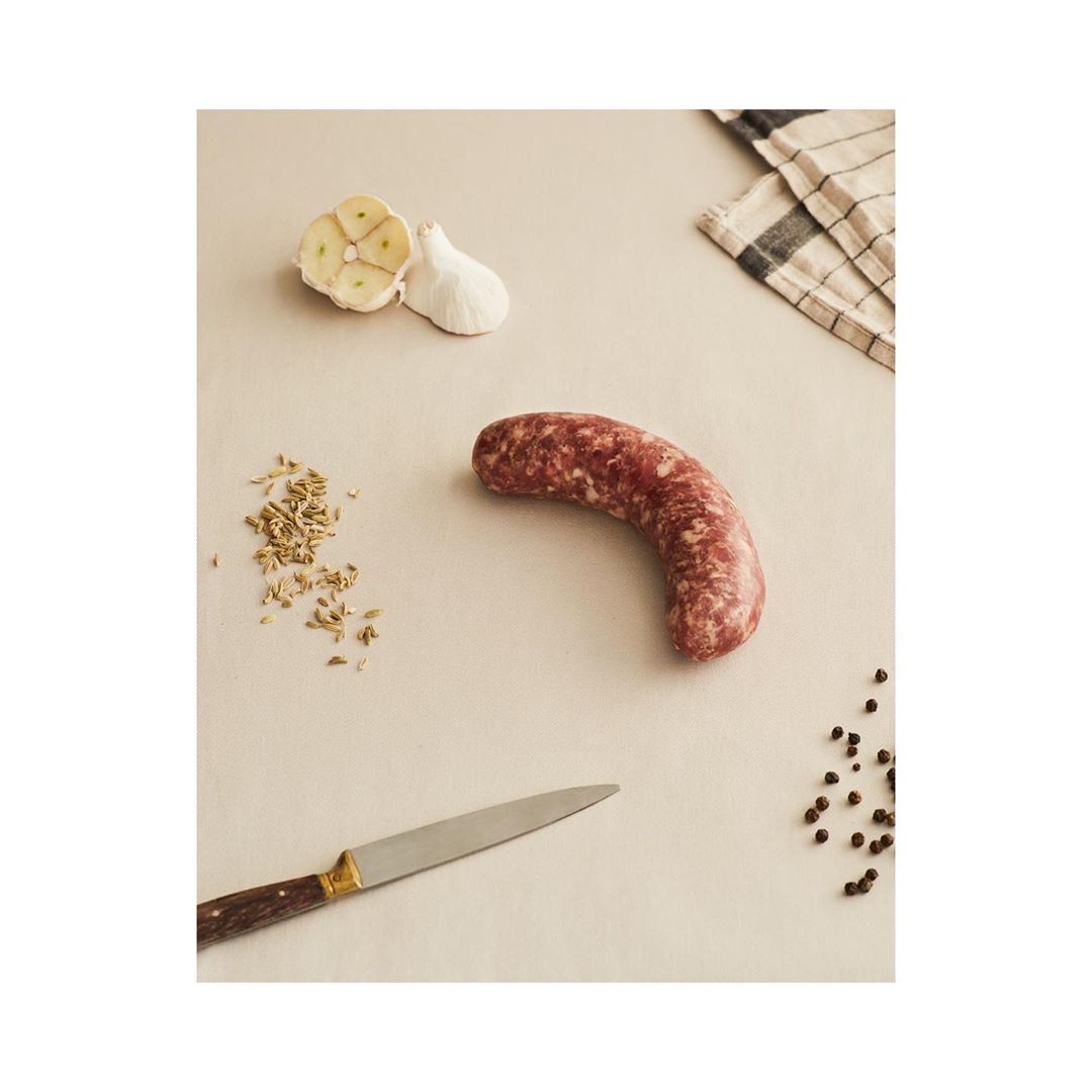 Overhead studio shot of raw sausage, garlic cloves, spices, peppercorns, and knife on a tan surface, perfect for culinary projects.