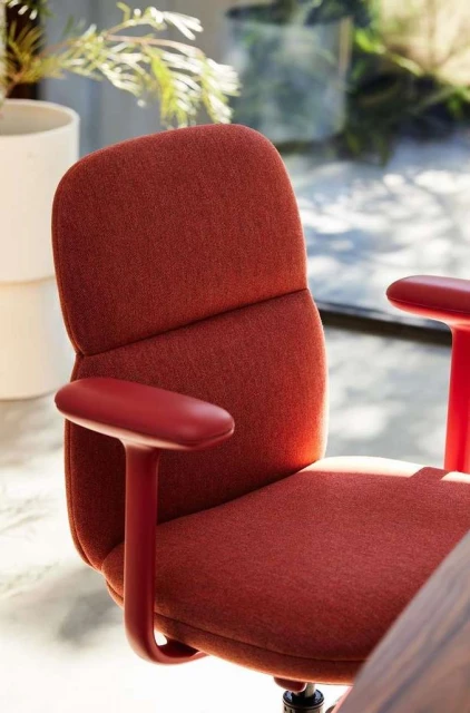 Close-up of a modern red office chair with a textured seat and back, set against a bright, blurred background with plants.