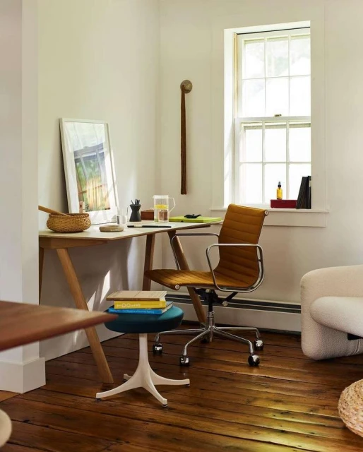 Office interior with a wooden desk, tan leather chair, stool with books, and natural light from a window, creating a cozy workspace.