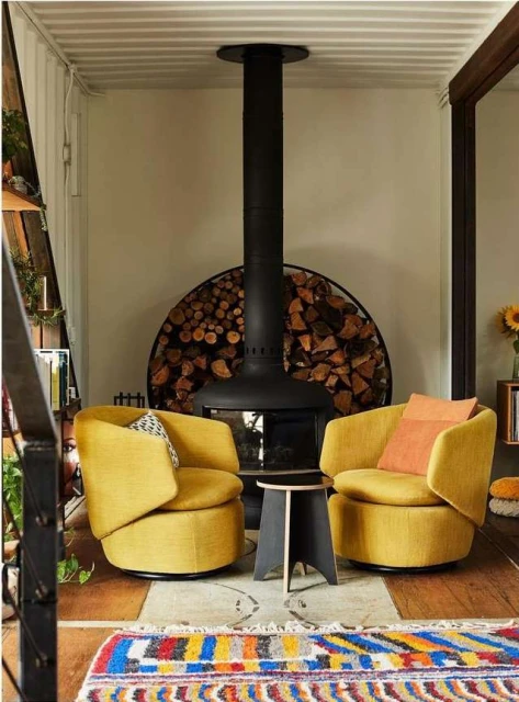 Living room interior with two yellow armchairs, a black fireplace filled with firewood, and a colourful patterned rug on the floor.