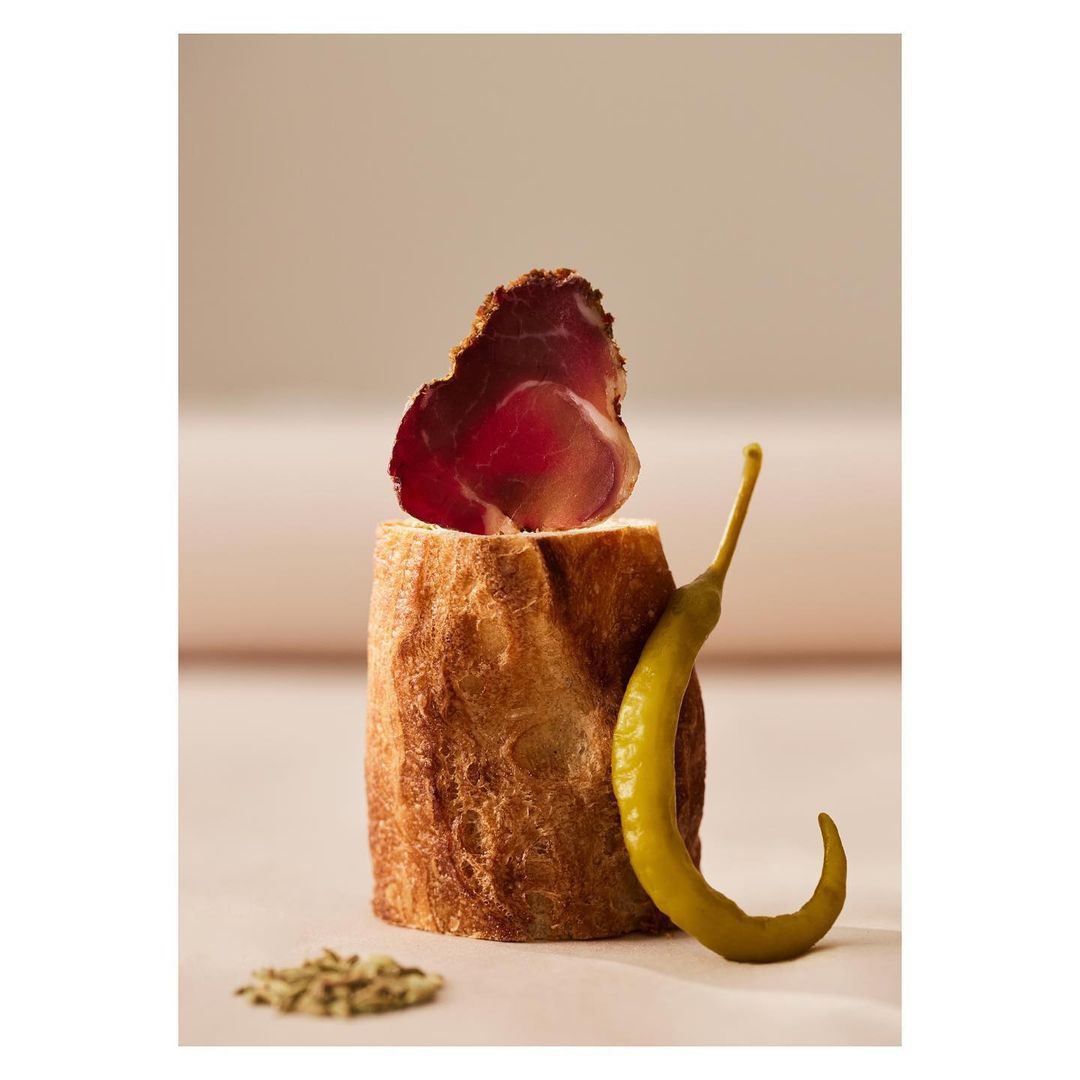 Rustic food still life featuring a slice of bresaola atop crusty bread, paired with a green pepper, and seasonings on a light surface.