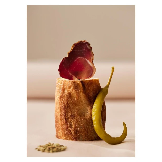 Rustic food still life featuring a slice of bresaola atop crusty bread, paired with a green pepper, and seasonings on a light surface.
