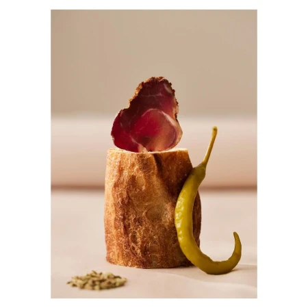 Rustic food still life featuring a slice of bresaola atop crusty bread, paired with a green pepper, and seasonings on a light surface.