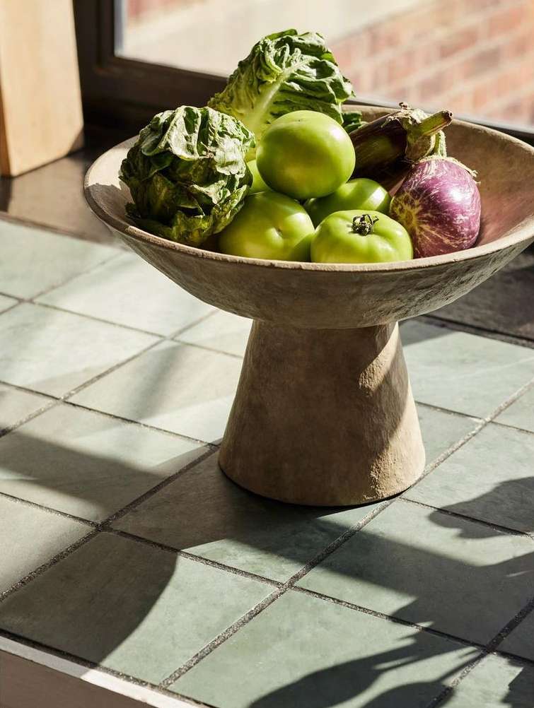 Still life of leafy greens, green tomatoes, and eggplants in a footed stone bowl, placed indoors on a tiled surface.
