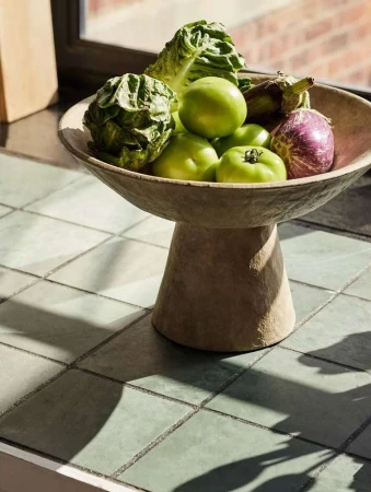 Still life of leafy greens, green tomatoes, and eggplants in a footed stone bowl, placed indoors on a tiled surface.
