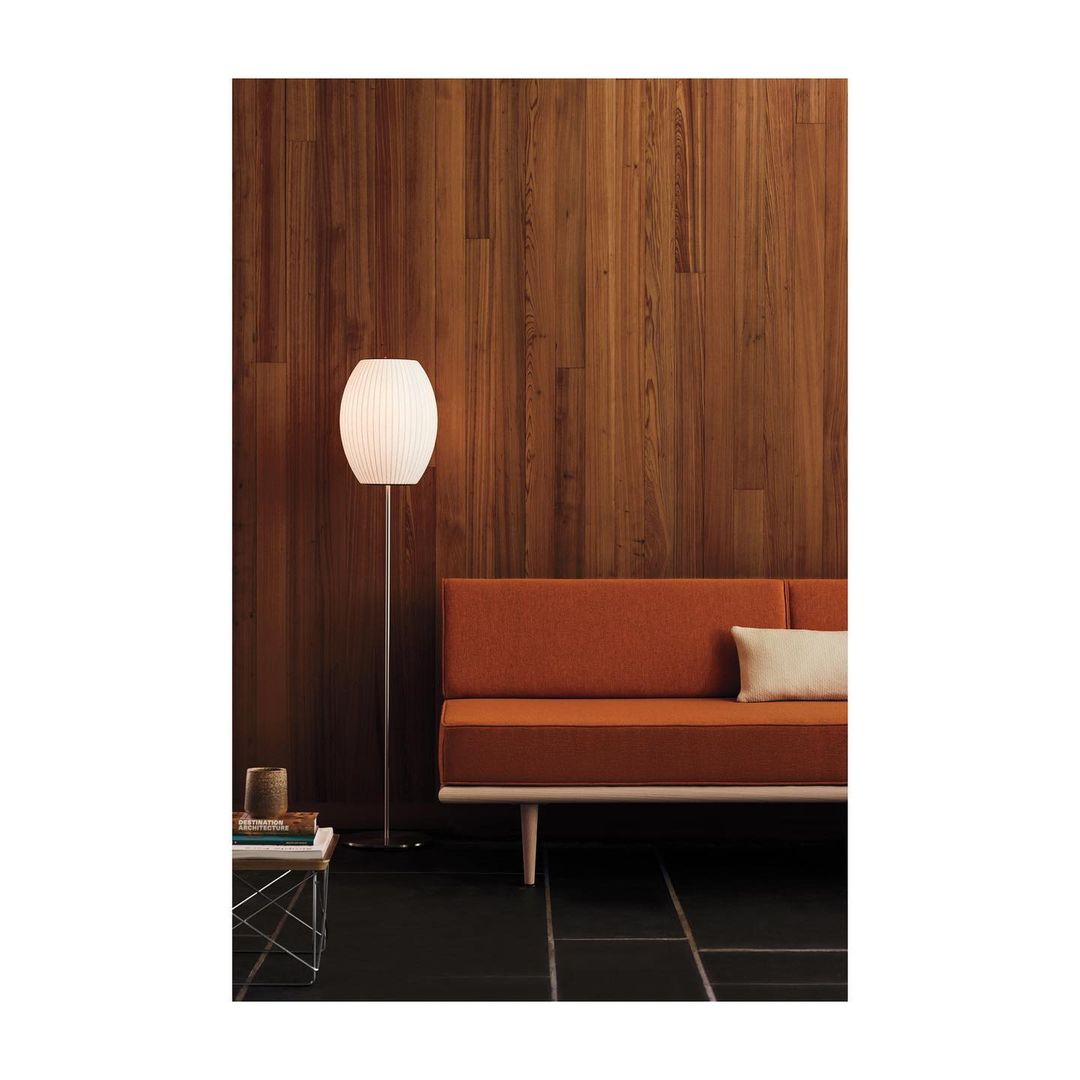 Modern interior featuring a floor lamp with a pleated shade next to an orange sofa against a wood-paneled wall and black tile floor.