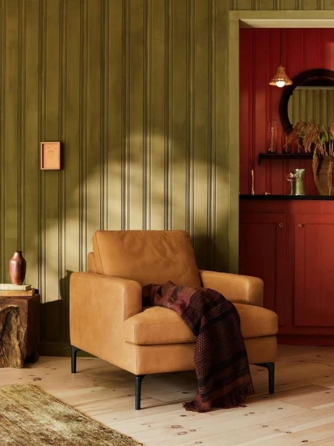 Living room interior featuring a caramel-colored leather armchair with a throw blanket, wooden floors, and green and red walls.