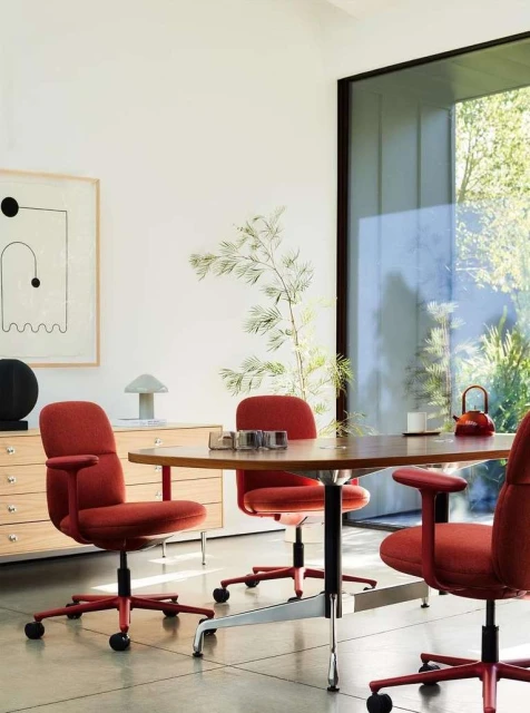 Interior shot of a conference room featuring red upholstered chairs around a wooden table, a sideboard, artwork, and natural light.