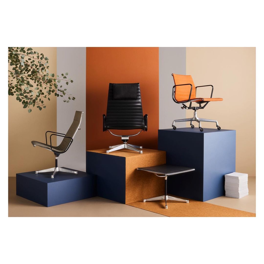 Three modern office chairs in black, orange, and grey are displayed on blue and brown platforms in a product studio shot.