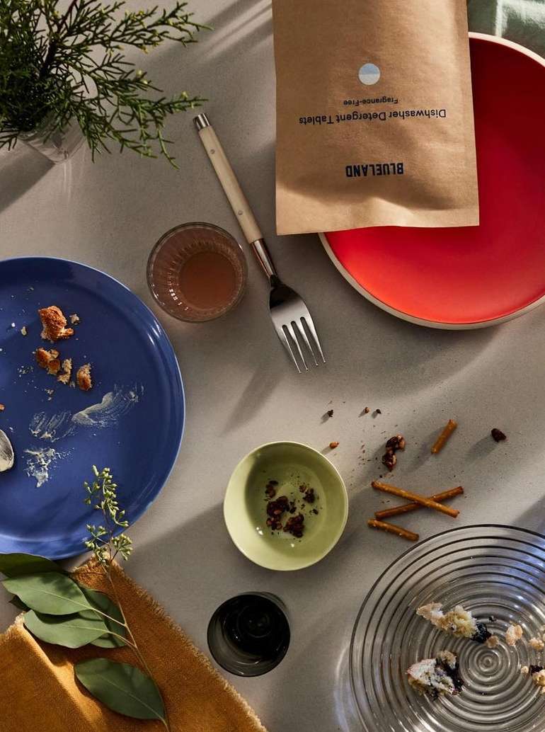 Overhead shot of an arrangement of used dishes, silverware, greenery, and Blueland dishwasher detergent tablets on a table.