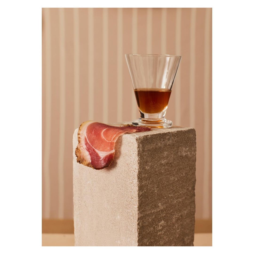 Still life photography featuring a glass of amber liquid, a slice of prosciutto, and a stone block against a striped background.