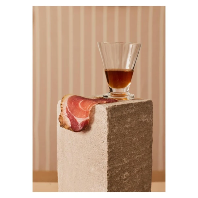 Still life photography featuring a glass of amber liquid, a slice of prosciutto, and a stone block against a striped background.
