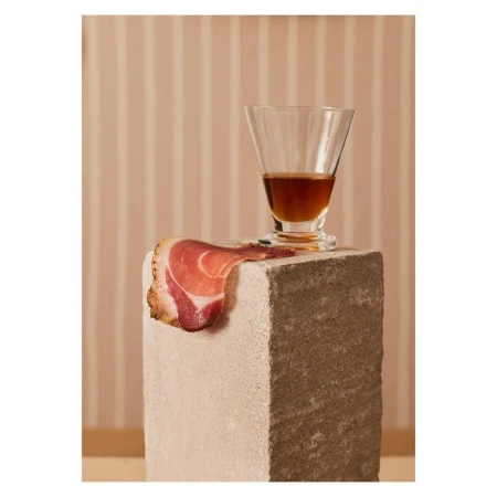 Still life photography featuring a glass of amber liquid, a slice of prosciutto, and a stone block against a striped background.