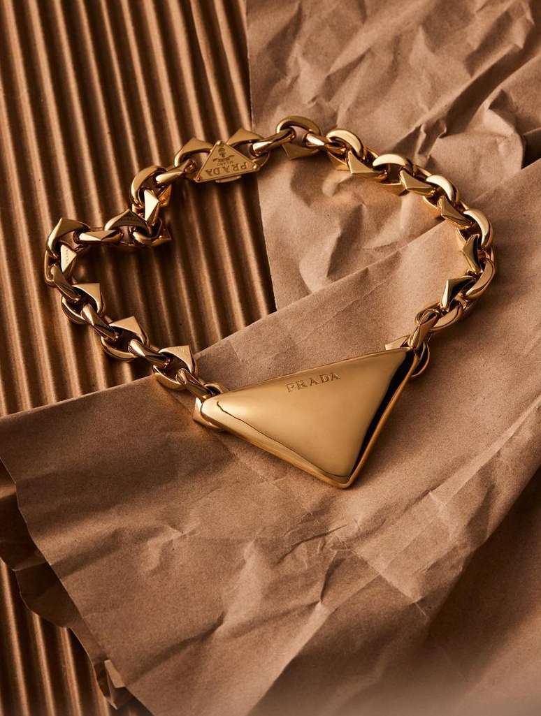 Close-up studio shot of a gold metal chain link Prada necklace with a triangle pendant on a textured brown paper background.