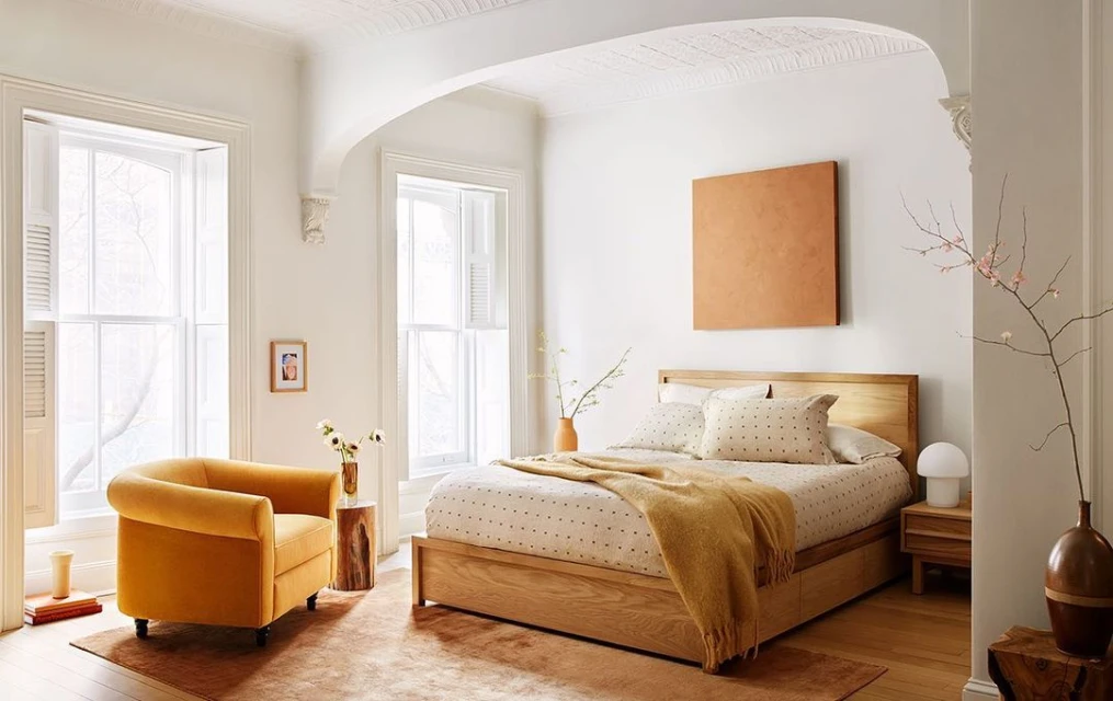 Bright bedroom interior with a wooden bed, patterned bedding, an armchair, a rug, and decor in natural light from windows.