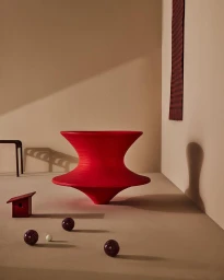 Vibrant red sculptural hourglass-shaped object resting on a neutral beige floor with small scattered spheres and a dark red wall hanging.
