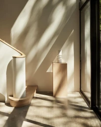 Minimalist interior with light casting diagonal shadows across a white wall, a wooden staircase, and a pedestal holding a sculptural object.