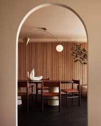 Interior shot framed through a white arched doorway, showing a dining table, chairs, modern pendant light, and slatted wood wall.