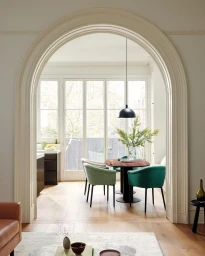 Interior dining space viewed through a large white architectural archway with green and grey upholstered chairs around a wooden table.