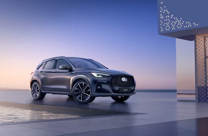 Sleek grey Infinity QX50 parked on concrete by reflective pool with modern architecture against clear blue and purple sky at dusk.