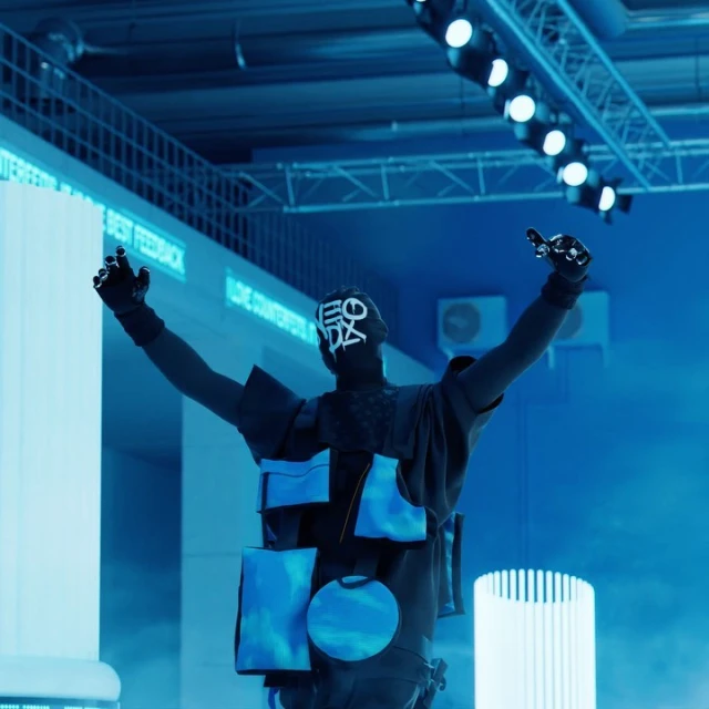 Back view of a person wearing an outfit with blue accents, raising their arms in a blue-lit space with overhead stage lighting.