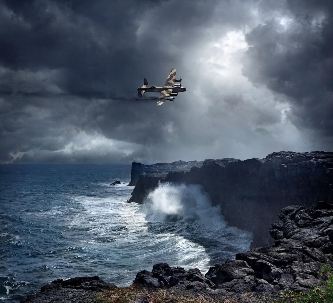 Vintage warplane flies over ocean cliffs with crashing waves and dark storm clouds in the sky, creating a dramatic aviation image.