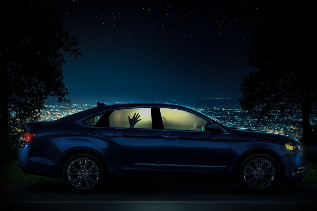 Dark blue sedan parked at night, with city lights glowing in the background and a hand visible inside the car window.