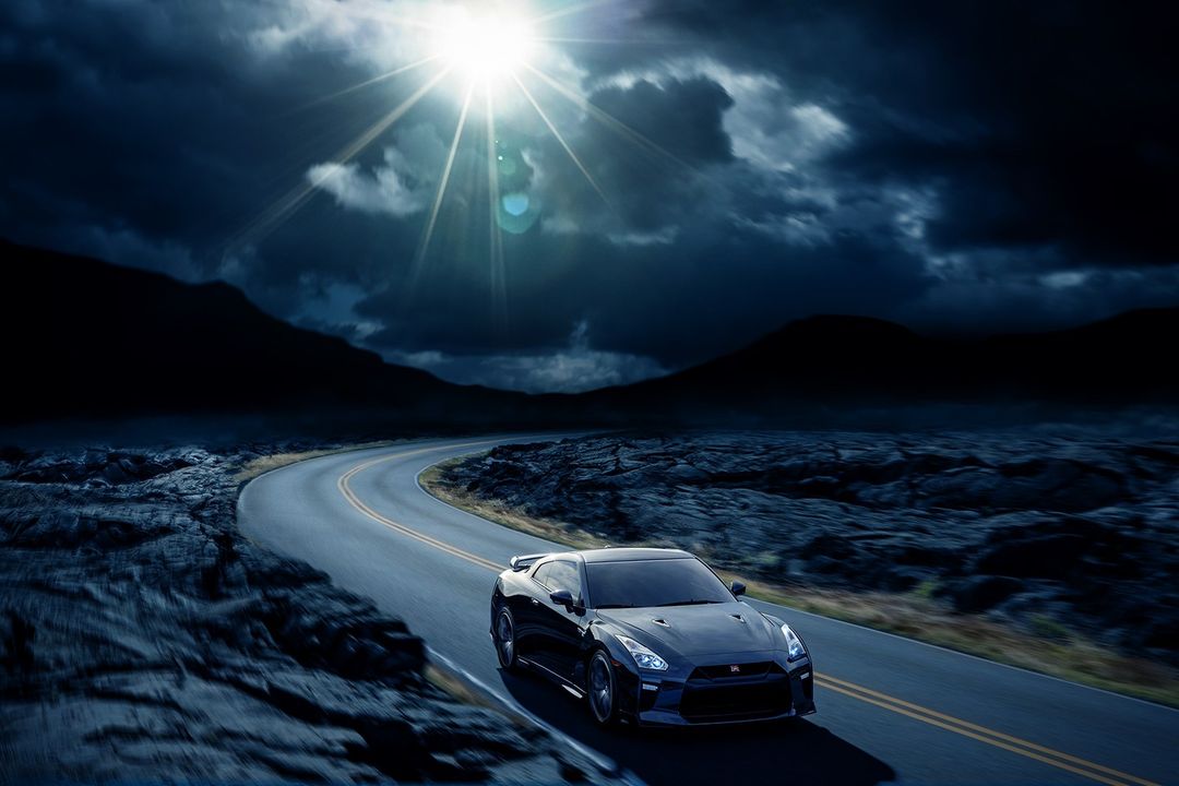 Sleek black sports car drives along a winding road under a bright, dramatic sky with dark clouds and distant silhouetted mountains.