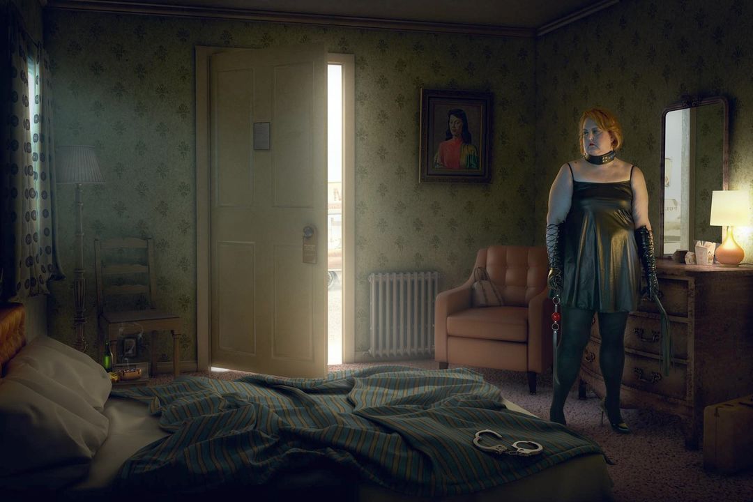 Adult female in black leather dress stands in bedroom holding a whip and leash with handcuffs visible on bed, surreal