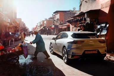 Silver electric SUV parked on a narrow sunlit street while a man pushes a laden cart past it in a historic market
