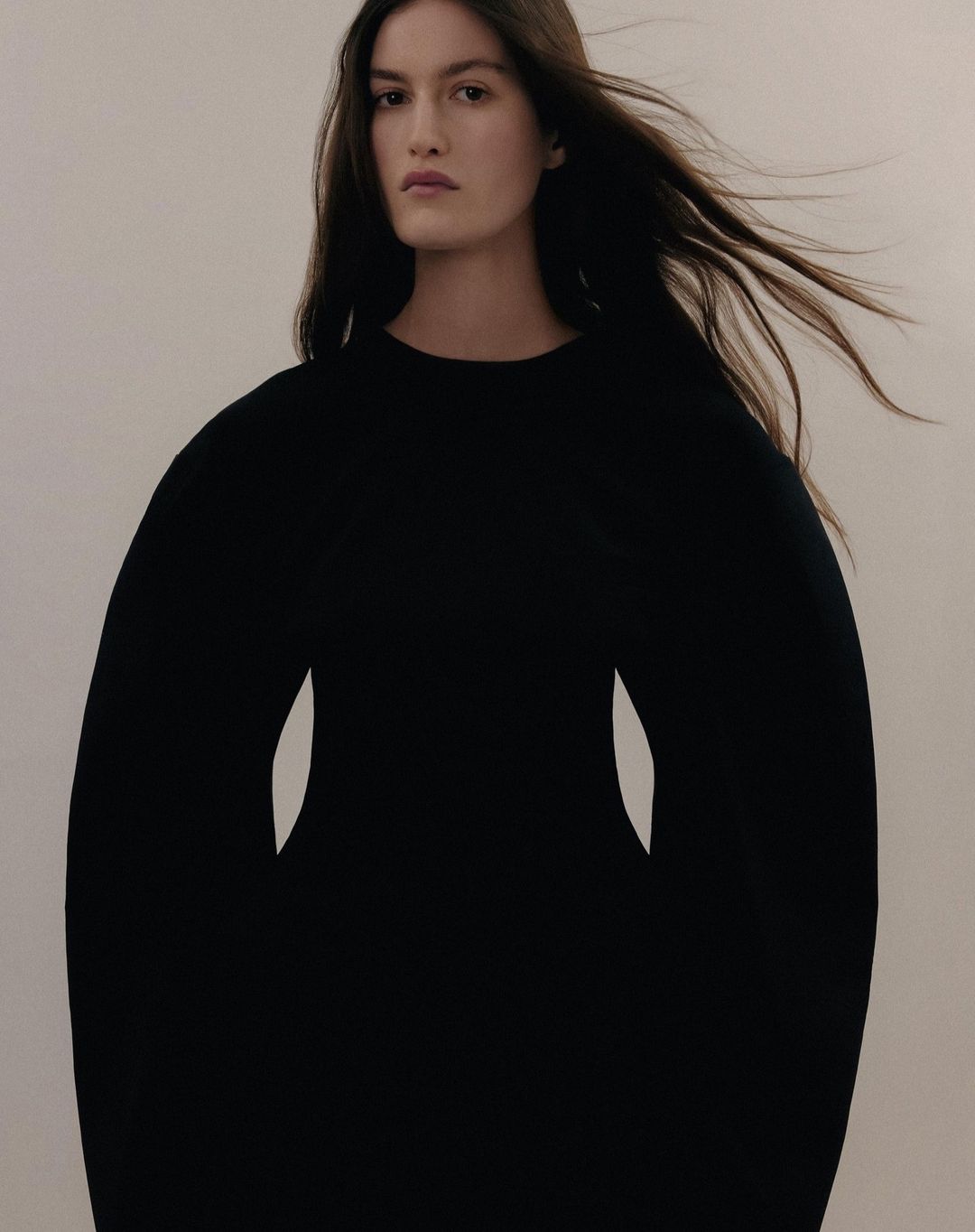 Portrait of a woman with long dark hair wearing a black long-sleeve dress against a neutral pale background.