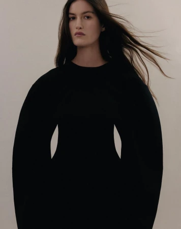 Portrait of a woman with long dark hair wearing a black long-sleeve dress against a neutral pale background.