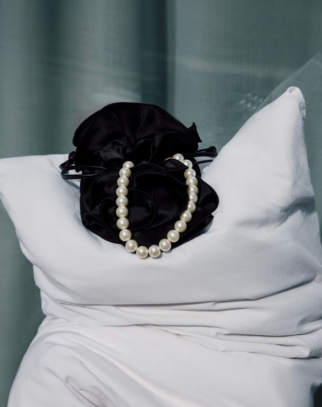 Elegant studio still life featuring a black satin drawstring bag and a pearl necklace resting on a soft white pillow and drapery.