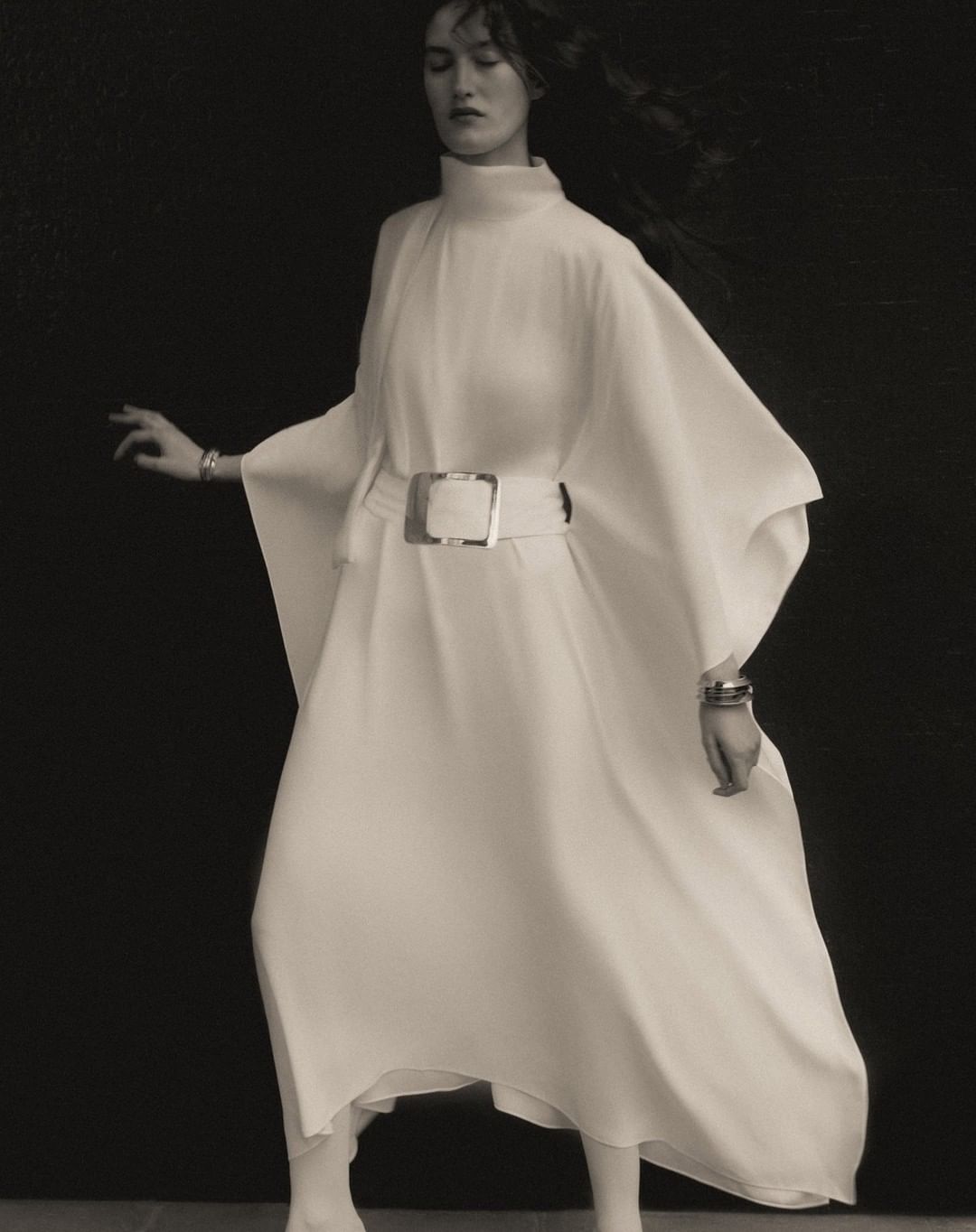 Monochrome fashion shot of a slender adult female model in a flowing white high-necked gown with a wide belt against a dark backdrop.