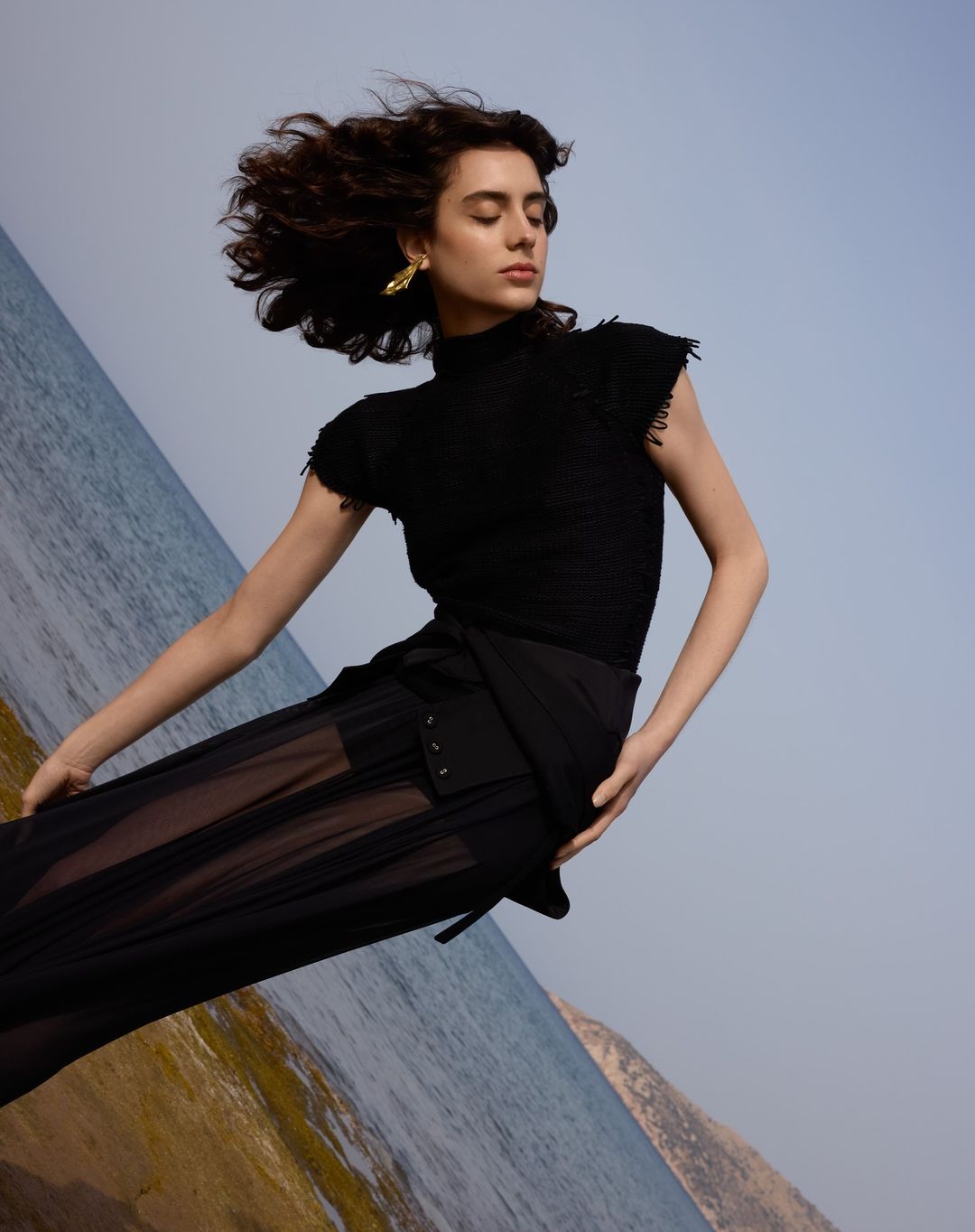 A woman in a black top and sheer black skirt poses in front of an angled ocean and sky, wearing gold earrings and with wind-blown hair.