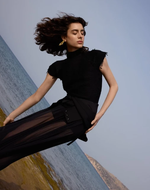 A woman in a black top and sheer black skirt poses in front of an angled ocean and sky, wearing gold earrings and with wind-blown hair.