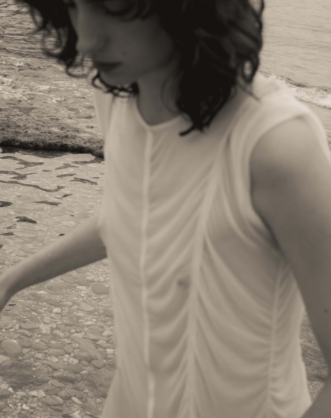 Woman in white dress on a rocky beach with waves in the background, creating a serene and muted coastal scene. Fashion photography.